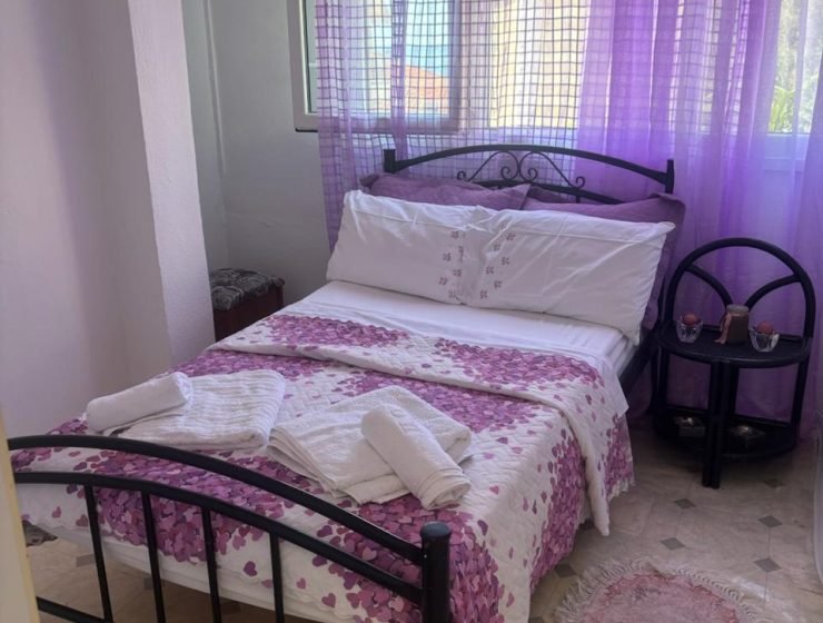Bedroom with lilac decor: sheer purple curtains, a metal-framed bed, white bedding with a pink floral quilt, and folded towels on the bed.