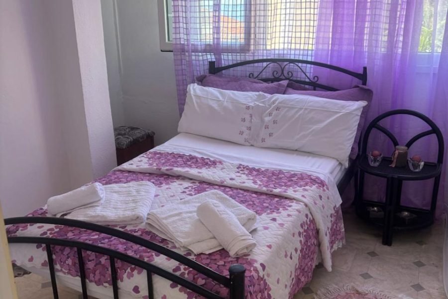 Bedroom with lilac decor: sheer purple curtains, a metal-framed bed, white bedding with a pink floral quilt, and folded towels on the bed.
