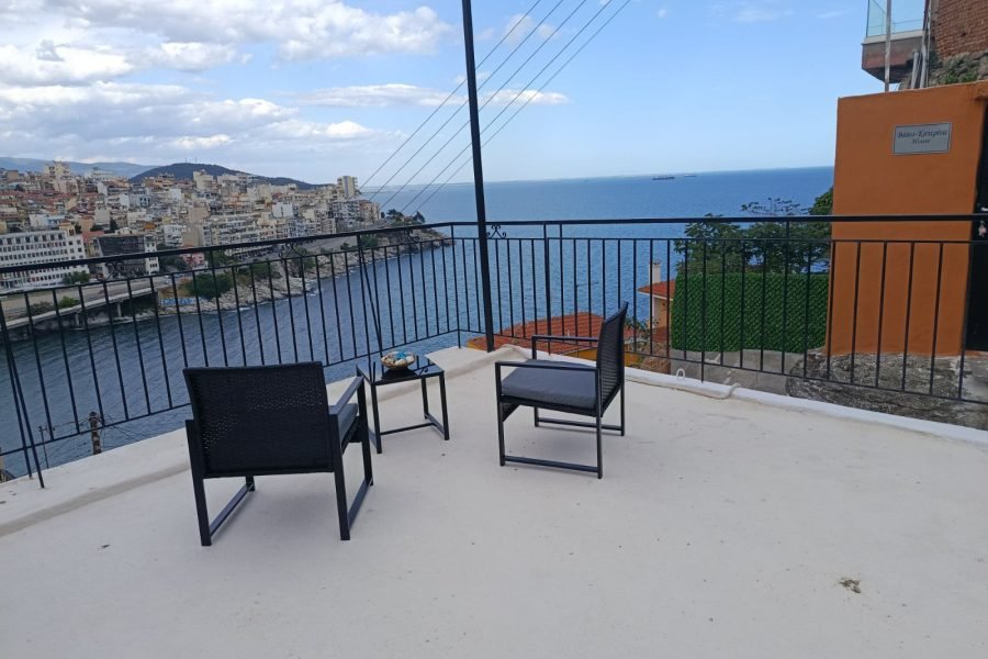 Terrace seating with two black chairs and a small table overlooking a coastal town and blue sea.