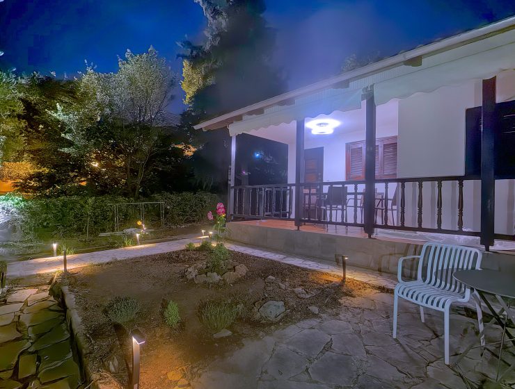 Nighttime view of a white house with a covered patio, railing, and garden lights amid trees and rocks.