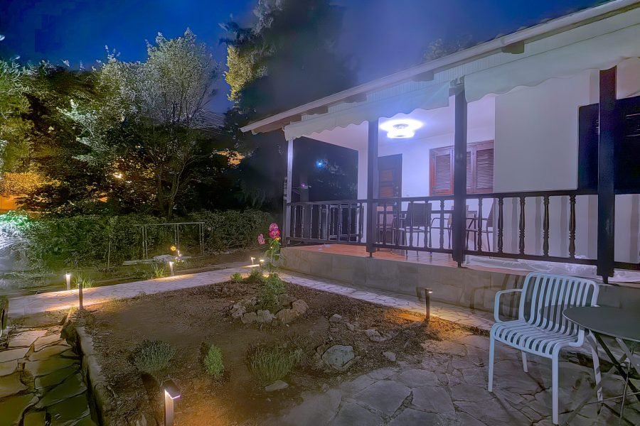 Nighttime view of a white house with a covered patio, railing, and garden lights amid trees and rocks.